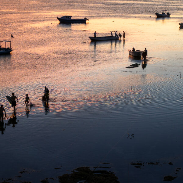 Surfers and local fishermen walking through shallow water among boats at sunset in a fine art coastal lifestyle photograph.