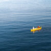 Two people seated in a small yellow boat floating on calm blue ocean water in a fine art ocean lifestyle photograph.