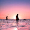 Surf lifestyle photograph of silhouetted surfers sitting in the ocean at sunset under a pink and purple sky.