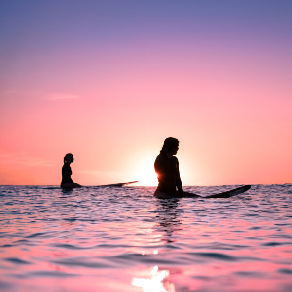 Surf lifestyle photograph of silhouetted surfers sitting in the ocean at sunset under a pink and purple sky.
