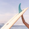 Close-up surf lifestyle photography showing a surfer holding a surfboard with a single fin against the ocean