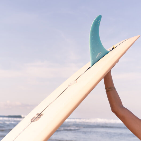 Close-up surf lifestyle photography showing a surfer holding a surfboard with a single fin against the ocean