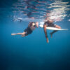 Two surfers floating underwater beside their boards in clear blue ocean water in a fine art surf lifestyle photograph.
