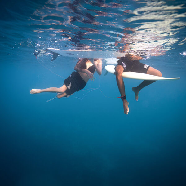 Two surfers floating underwater beside their boards in clear blue ocean water in a fine art surf lifestyle photograph.