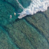 Aerial surf photography showing surfers paddling across a reef as a wave breaks overhead