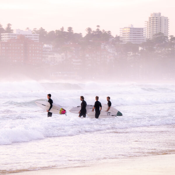 Four surfers walking into the ocean at sunrise with boards under their arms, soft pastel light over a misty Manly Beach.