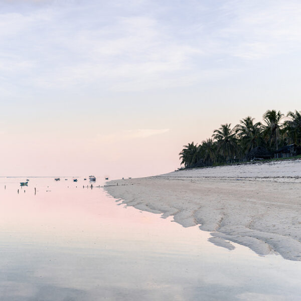 Pastel sunrise over a quiet tropical beach with palm trees and anchored boats reflected in calm water.