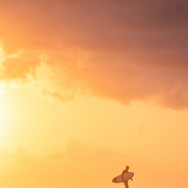 Sunset surf photography showing a surfer walking along the shoreline beneath a moody yet golden sky