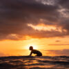 Sunset surf photography showing a surfer silhouetted against a moody, stormy sky while sitting on a board at dusk