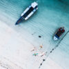 Surf lifestyle photography showing surfers walking across shallow turquoise water beside anchored boats