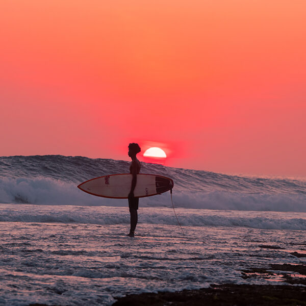 Surfer standing in shallow water at sunset, holding a surfboard as waves roll in behind, silhouetted against a coral-pink sky.